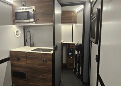 Interior view of the CORE 6515 Trailhead with closet door open and looking towards the kitchenette with upper microwave, 2 burner cooktop and small sink