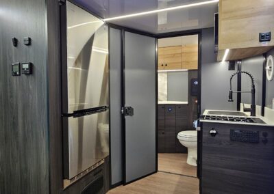 Interior view of a CORE RV kitchen showing a large stainless steel refrigerator, a look through the bathroom, and kitchen cooktop and sink.